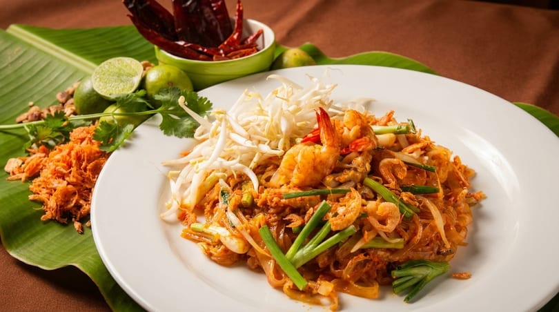 A plate of shrimp pad Thai with bean sprouts, limes, and dried chilies on a banana leaf.