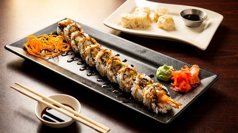 A long black plate of sushi rolls with sauce, wasabi, ginger, and carrot strings, with dumplings in the background.