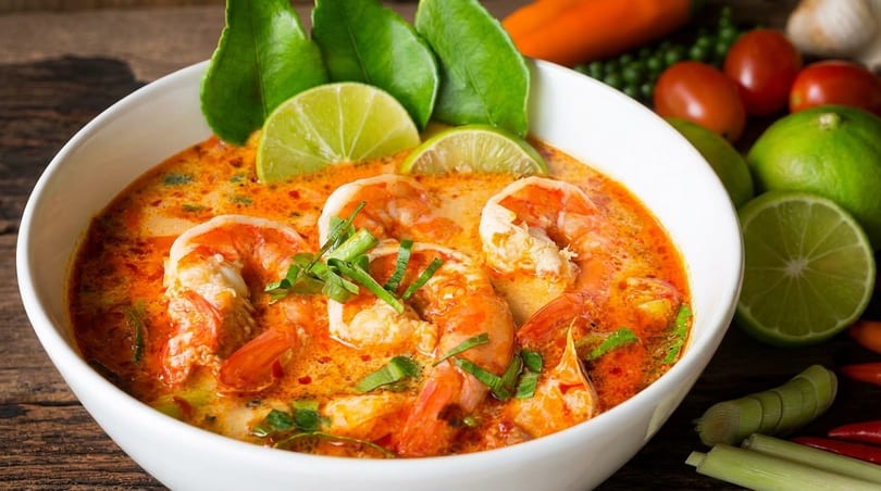 A white bowl of Tom Yum Goong soup with shrimp, lime, and herbs on a wooden table.